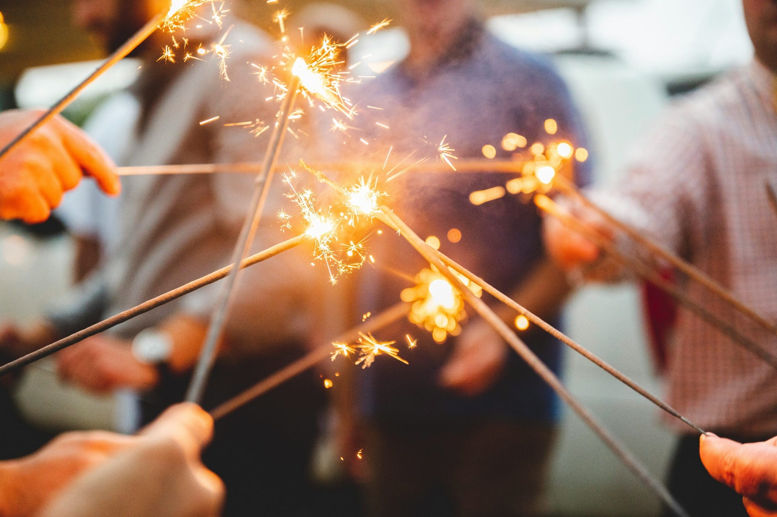 Several people hold lit sparklers close together, creating bright sparks, with blurred figures and a vehicle in the background.
