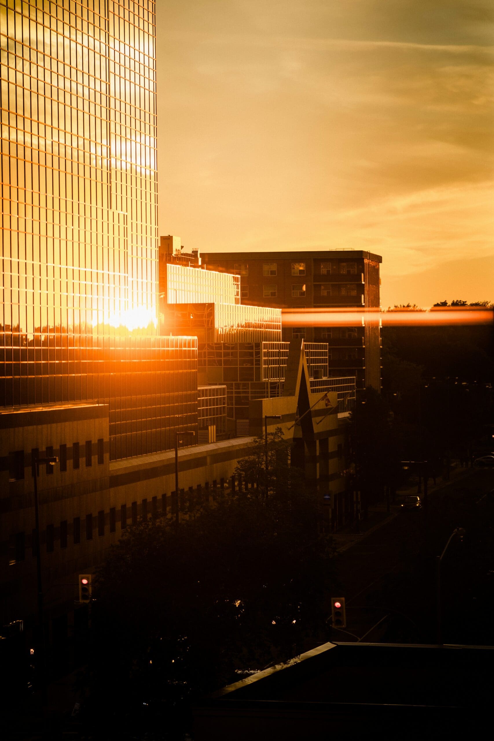 Sunset reflects off the windows of a modern city building, casting a warm, golden light over nearby structures and the street below.